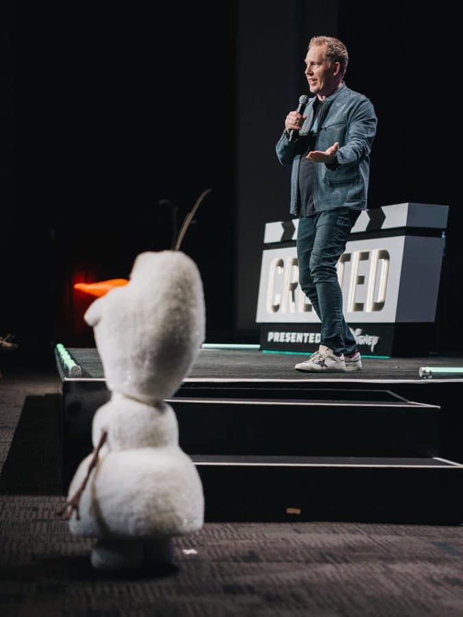 Speaker on a dark stage holding a microphone gesturing as he talks with a white stuffed figure in the foreground
