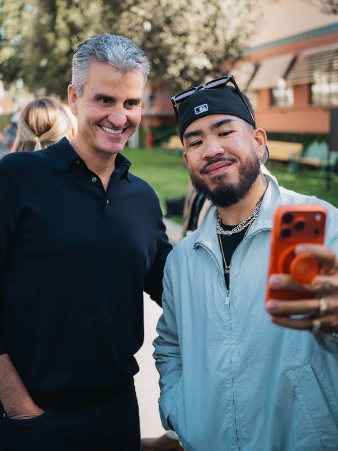 Two men smiling for a selfie outdoors one in a black polo and the other in a light blue jacket holding an orange phone to take the photo