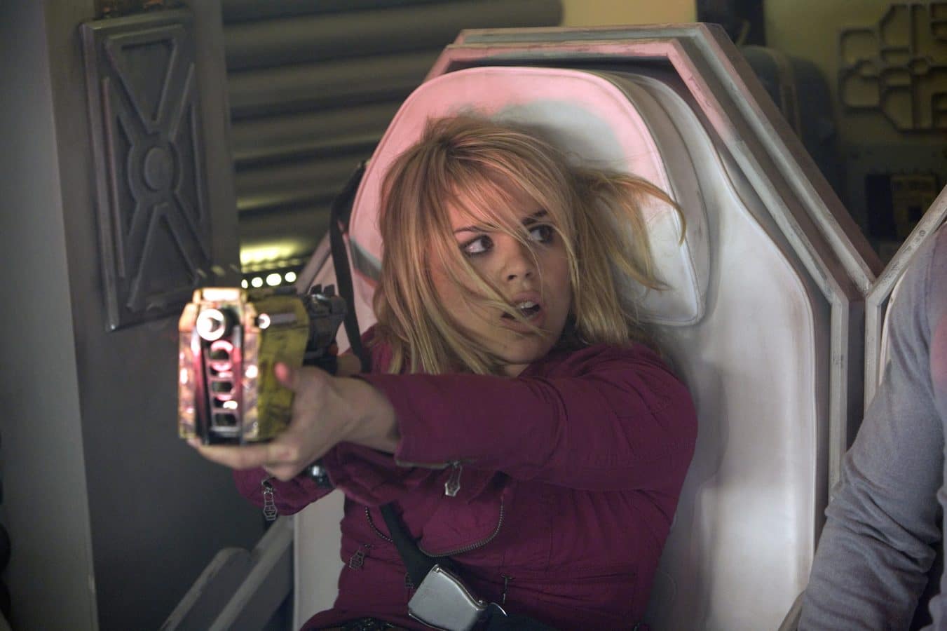 Woman in a burgundy jacket aiming a handheld weapon inside a sci fi pod or capsule