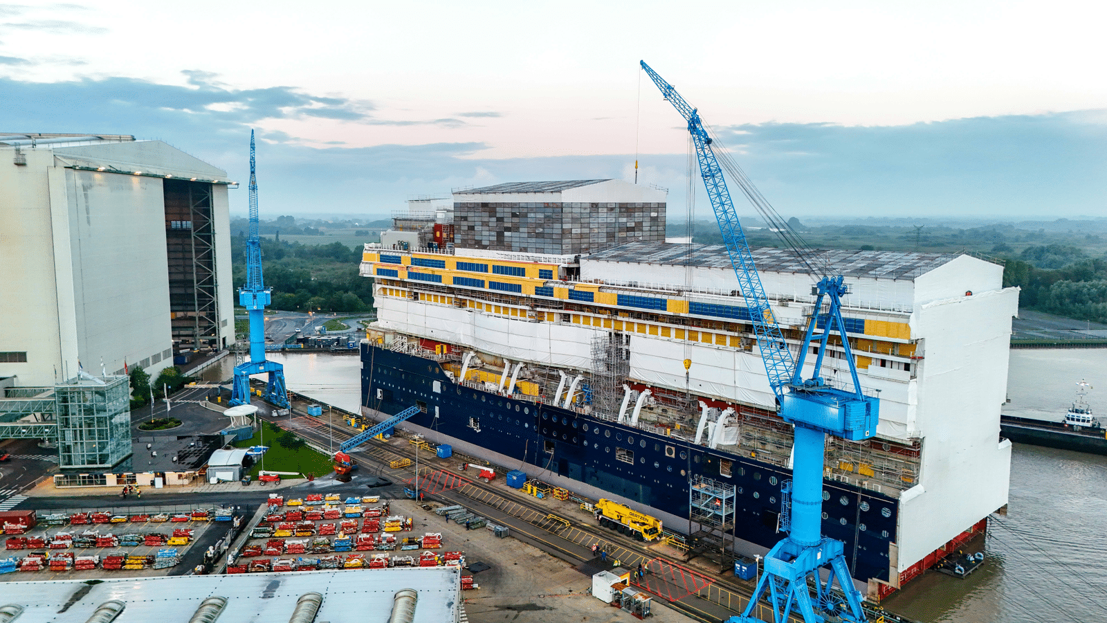Aerial view of a shipyard with large blue cranes a partially built vessel and industrial buildings by the water