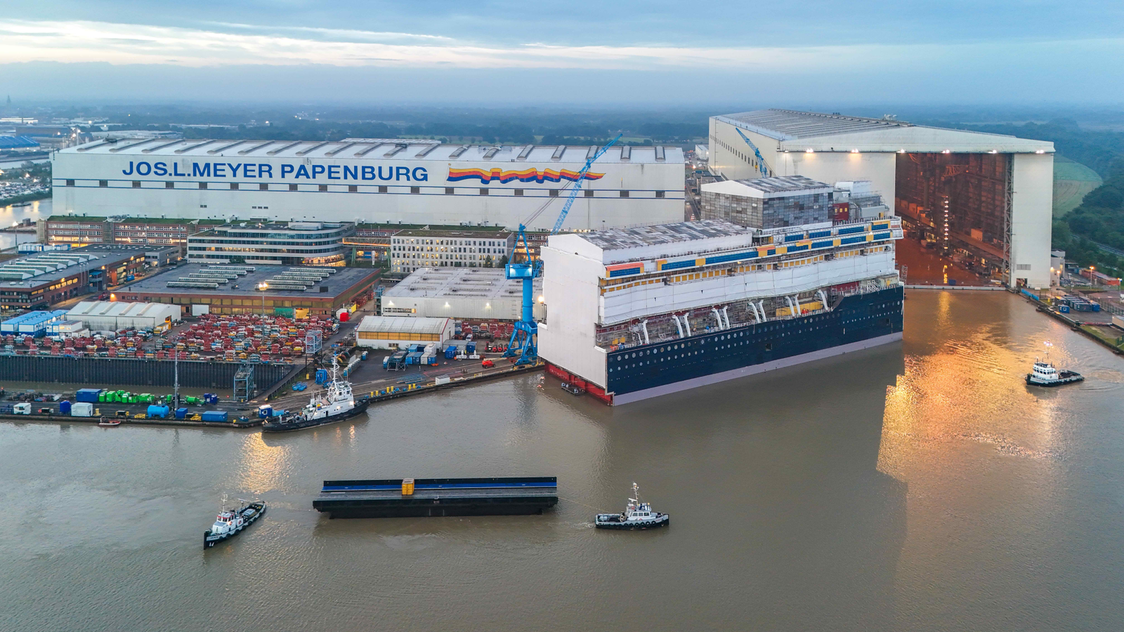 A large cruise ship under construction in a shipyard with cranes and industrial buildings nearby