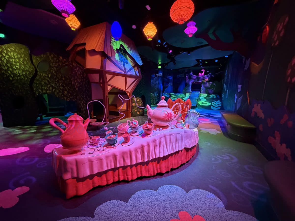 Colorful tea party setup in a neon lit fantasy room with a long pink table teapots cups and saucers below glowing lanterns