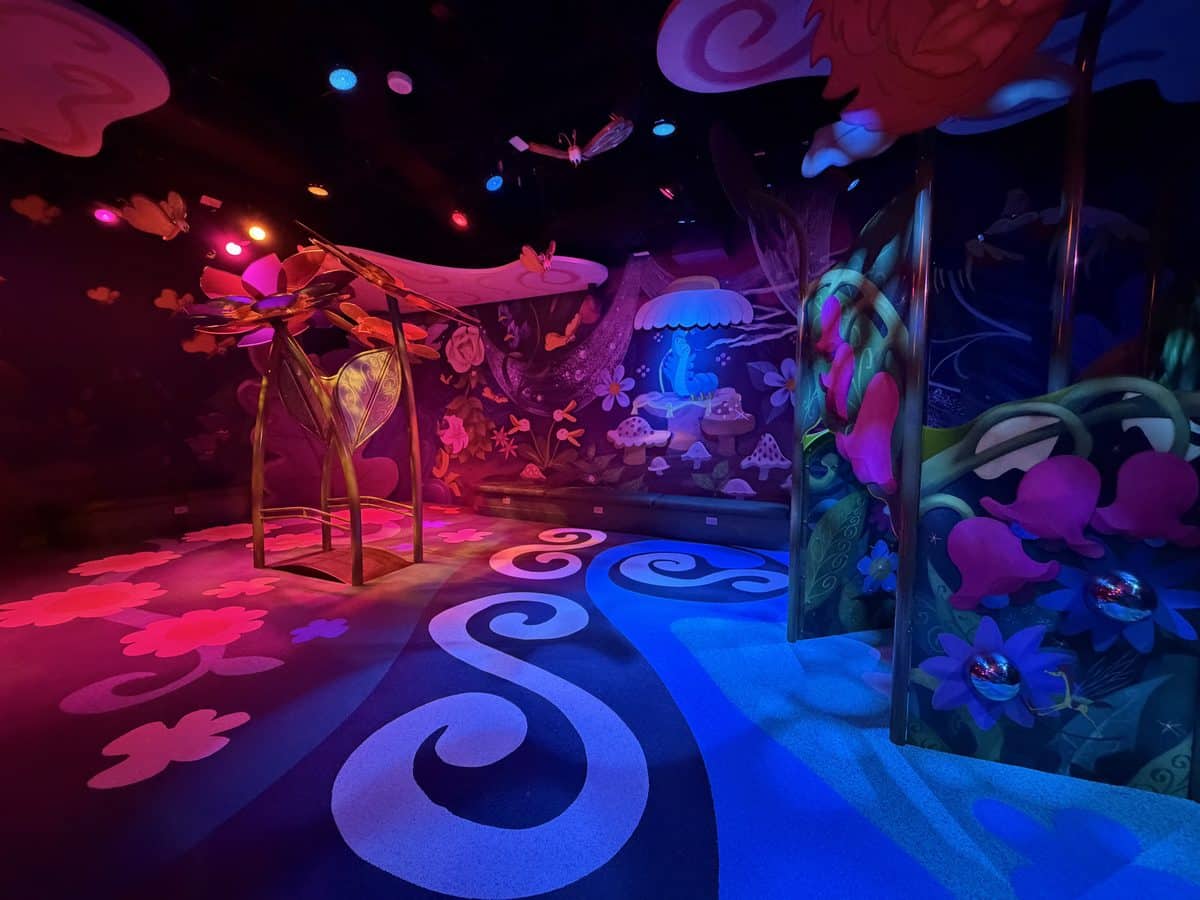 Colorful fantasy mural with oversized flowers mushrooms and swirl patterns illuminated by pink and blue lights in a dark room