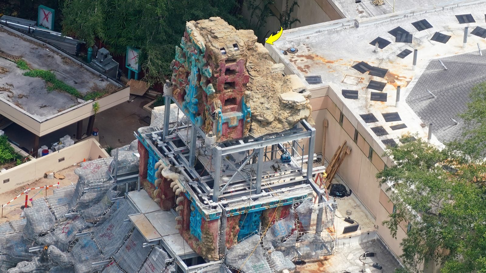 Construction site featuring a weathered multicolored rock sculpture on a metal framework surrounded by chain link fencing and rubble