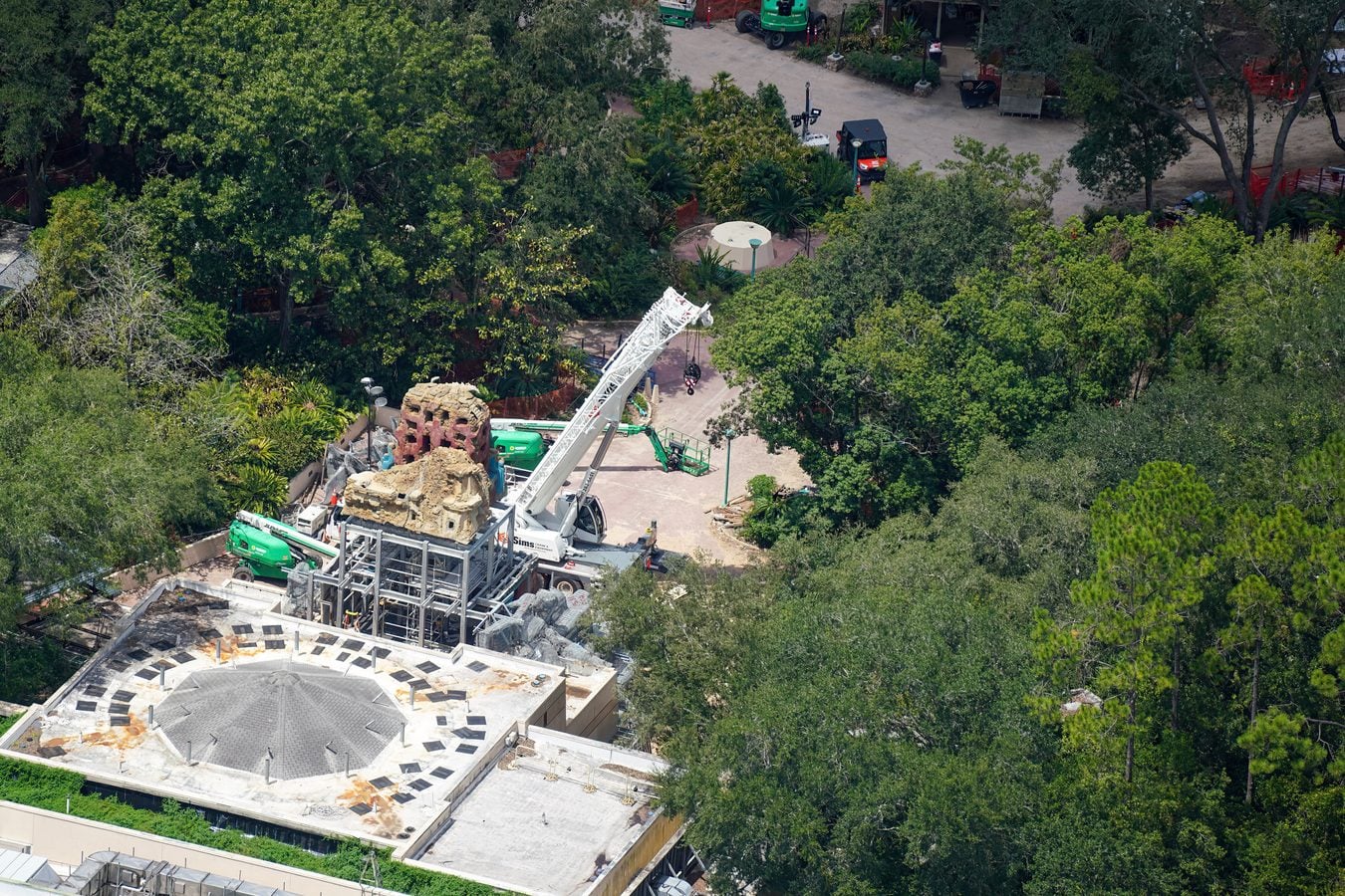 Aerial view of a construction site on a partially built building with a tall white crane and surrounding green trees nearby