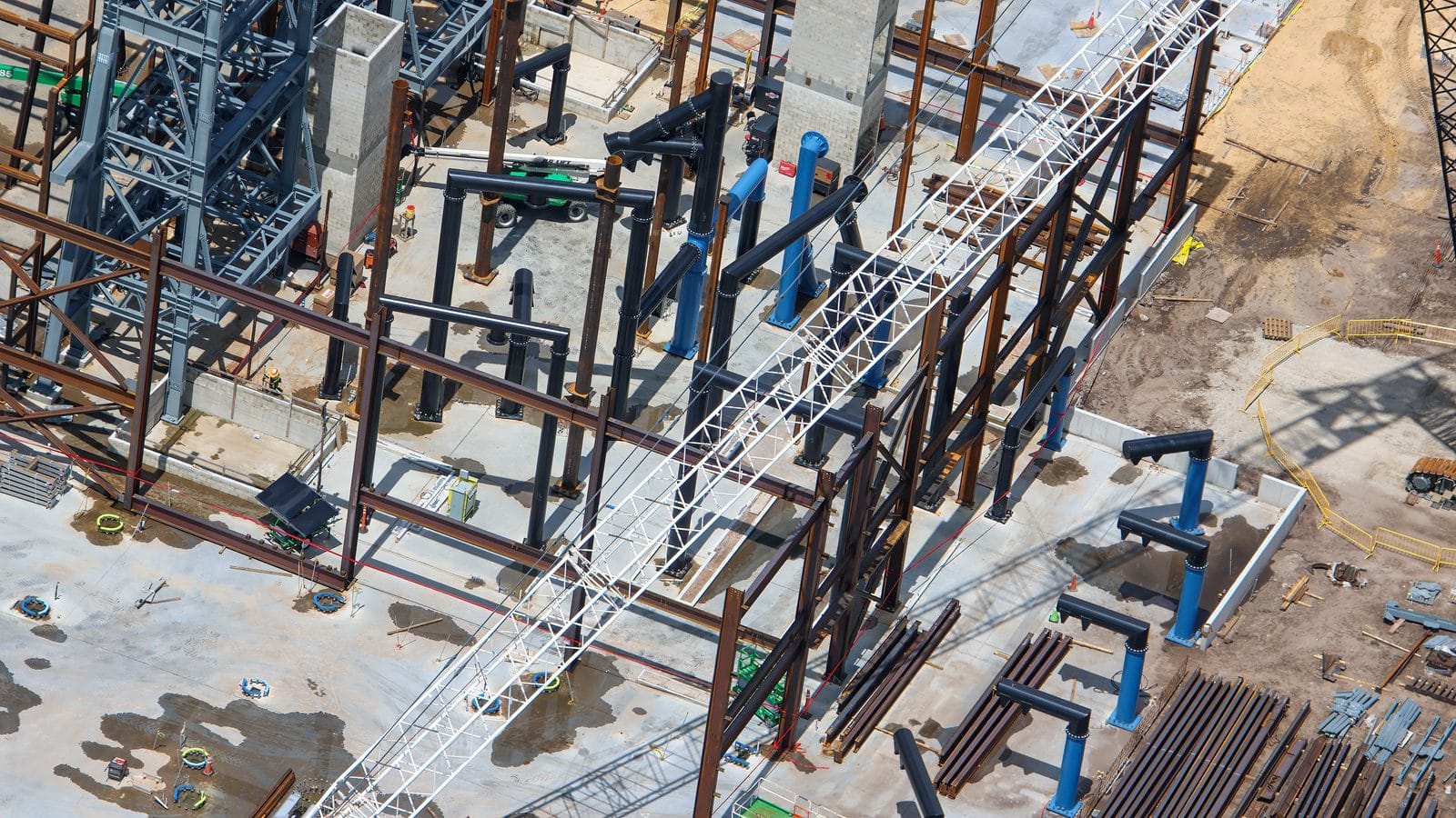 Aerial view of a construction site with steel framing blue pipes and a white lattice crane overhead from above ground level