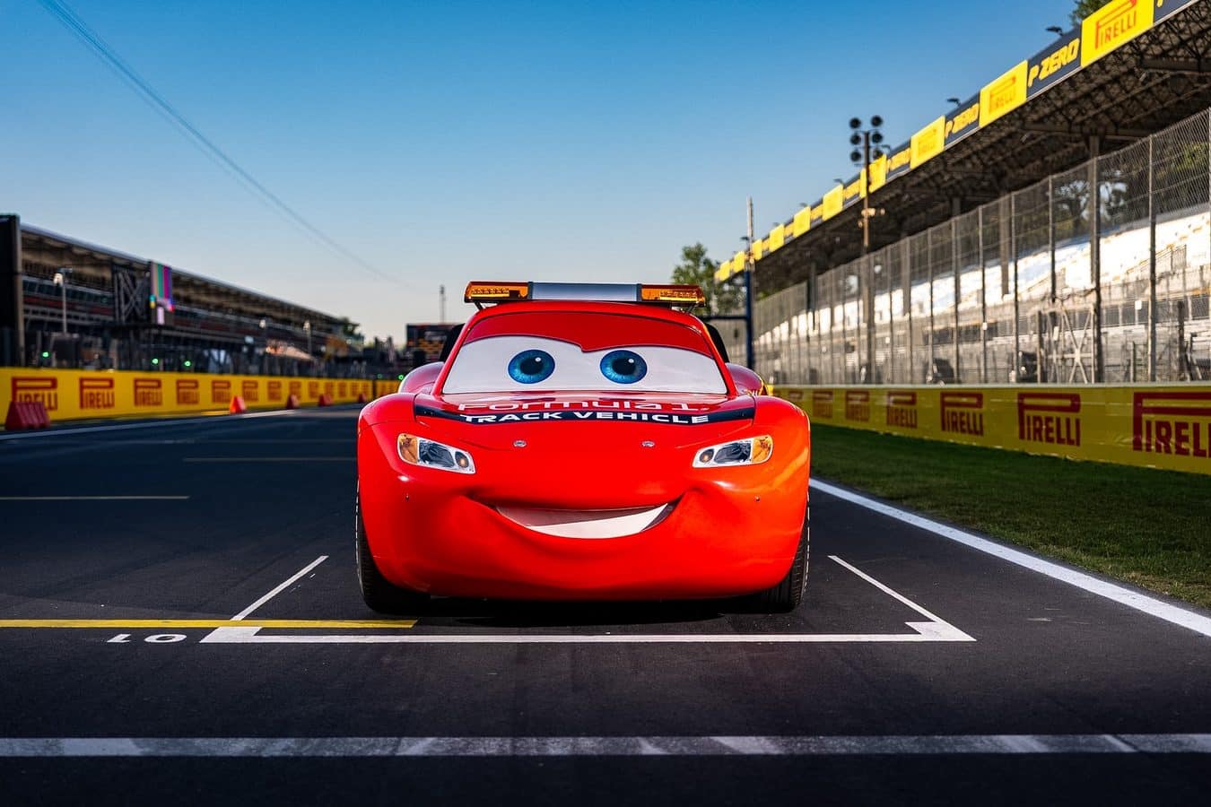 Front view of a red cartoon style race car with big blue eyes and a wide smile positioned at the starting line of a race track with yellow barriers in the background