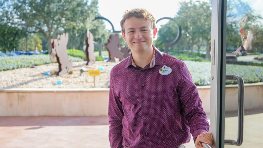 Smiling man in a maroon shirt with a name badge stands at a glass door entry outdoors in a courtyard park setting