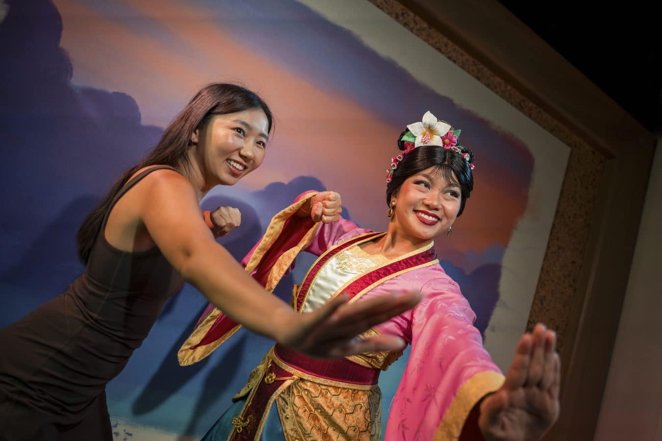 Two women dancing on stage one in a bright pink traditional costume with a floral headpiece smiling as they move together