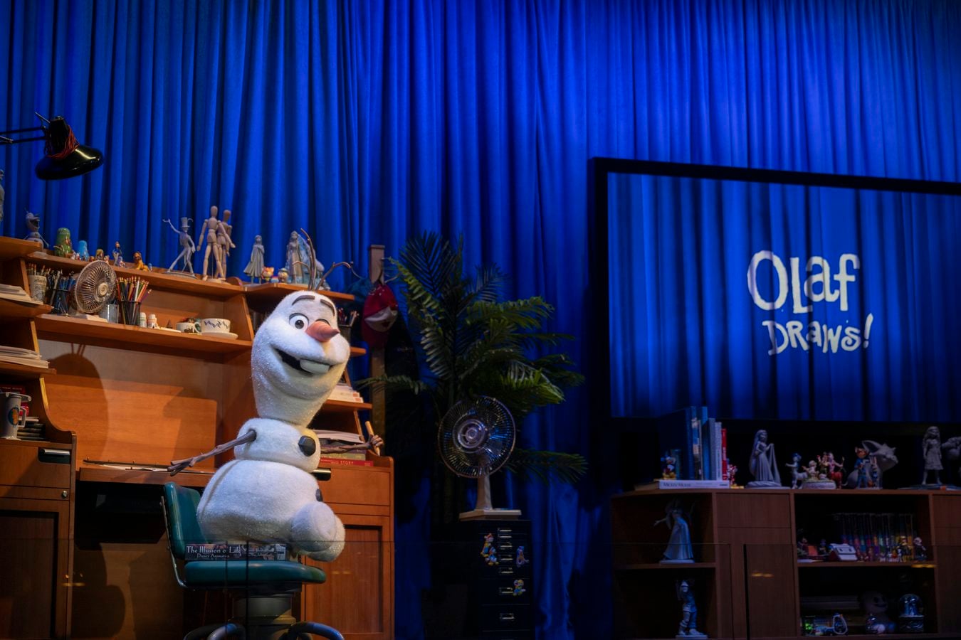 Life size Olaf puppet on a desk with art supplies figurines and shelves against a blue theater curtain backdrop and a sign that says Olaf Draws