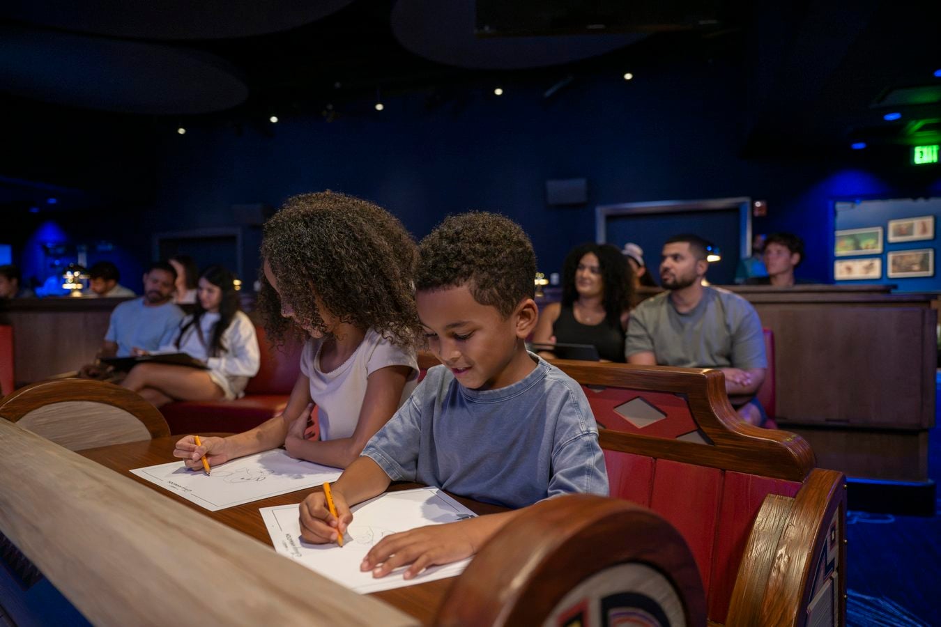 Two children in foreground coloring with pencils at a long wooden table in a dim blue lit room while other attendees sit in the background
