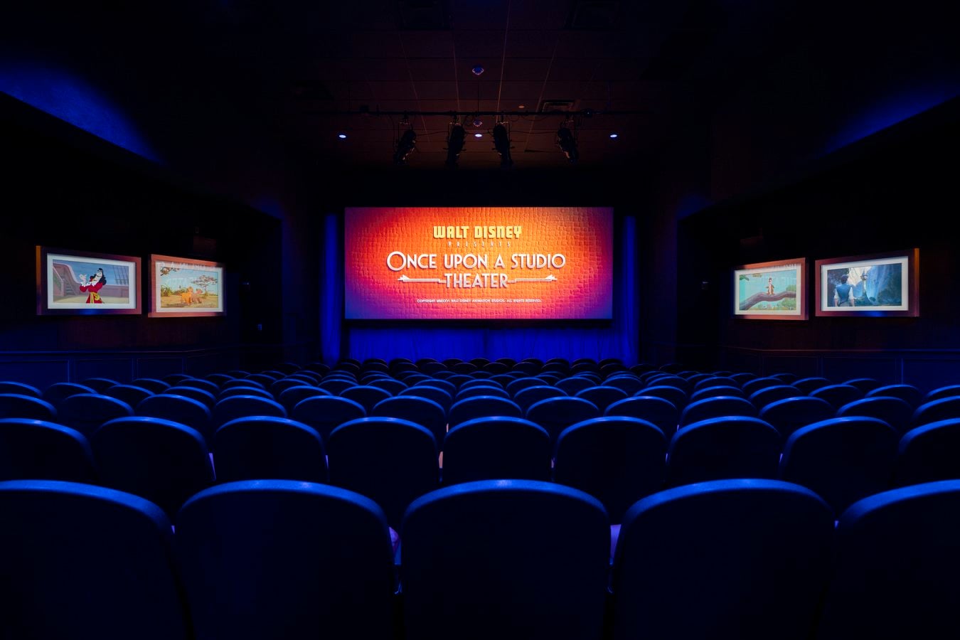 Dark theater with rows of blue seats facing a brightly lit orange stage backdrop reading Once Upon a Studio Theater and framed screens on the side walls