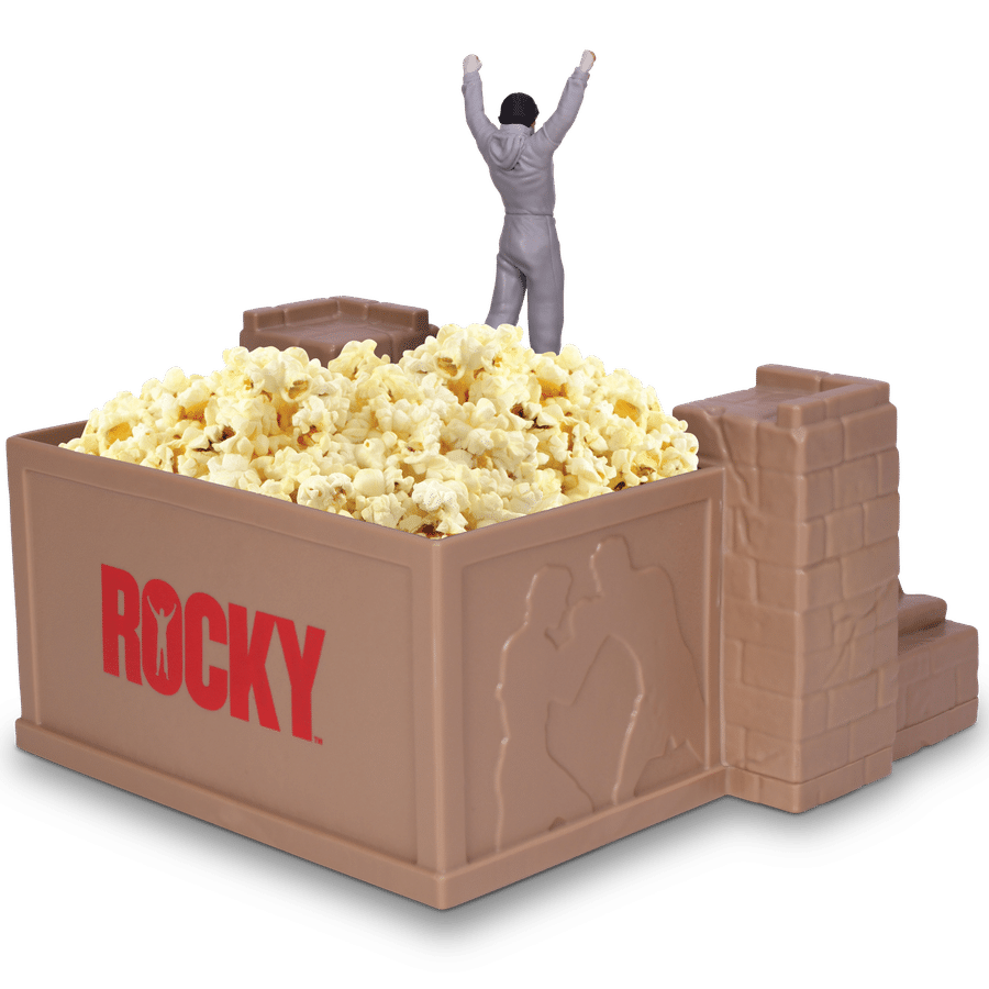 Gray figurine with arms raised stands in a large open box filled with popcorn and surrounded by chocolate bars suggesting a celebratory snack scene