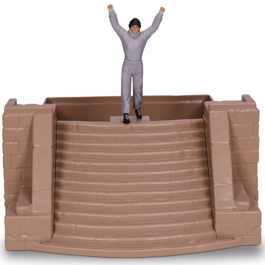 Person in a gray hoodie and beanie raises both arms while standing on a brown stair like platform Baseplate style blocks beneath him