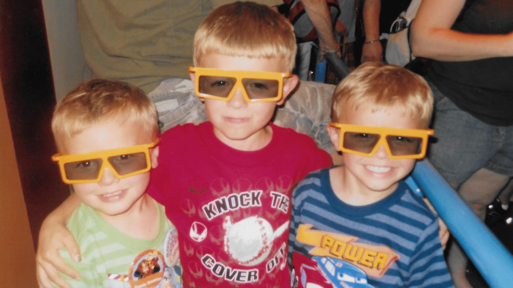 Three young boys with orange 3D glasses pose together arms around each other at an indoor event