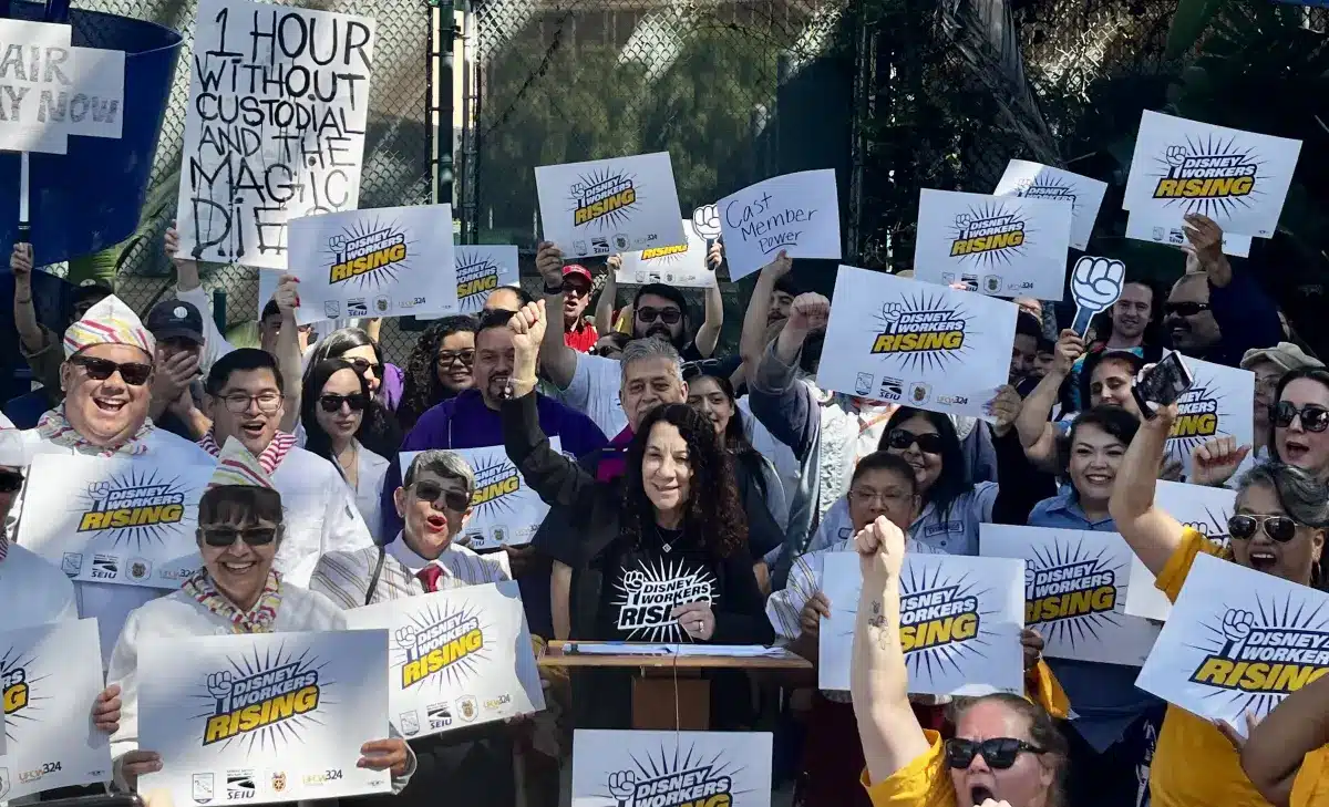 Crowd of diverse activists at a rally many holding signs reading Disney Workers Rising around a central speaker at a podium
