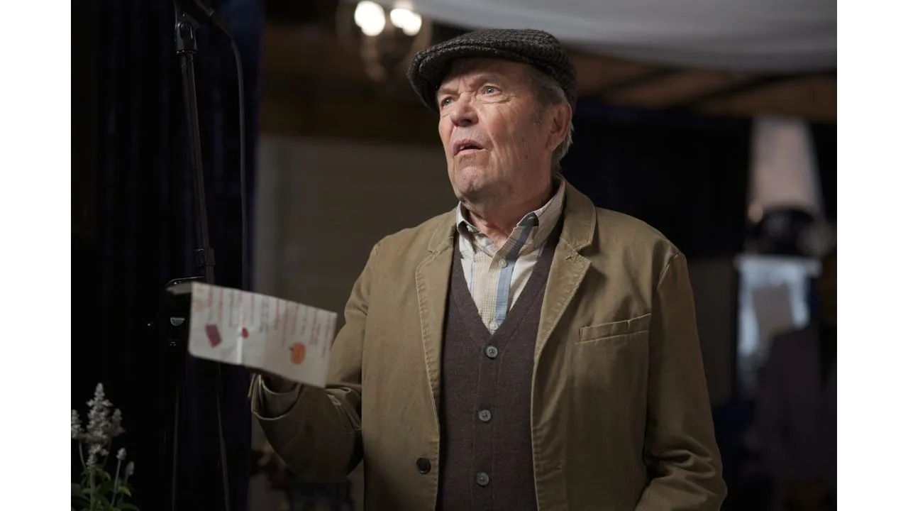fallplace2 Fantasy Land News Older man on a stage wearing a flat cap brown vest and beige blazer holding a folded program and looking thoughtful