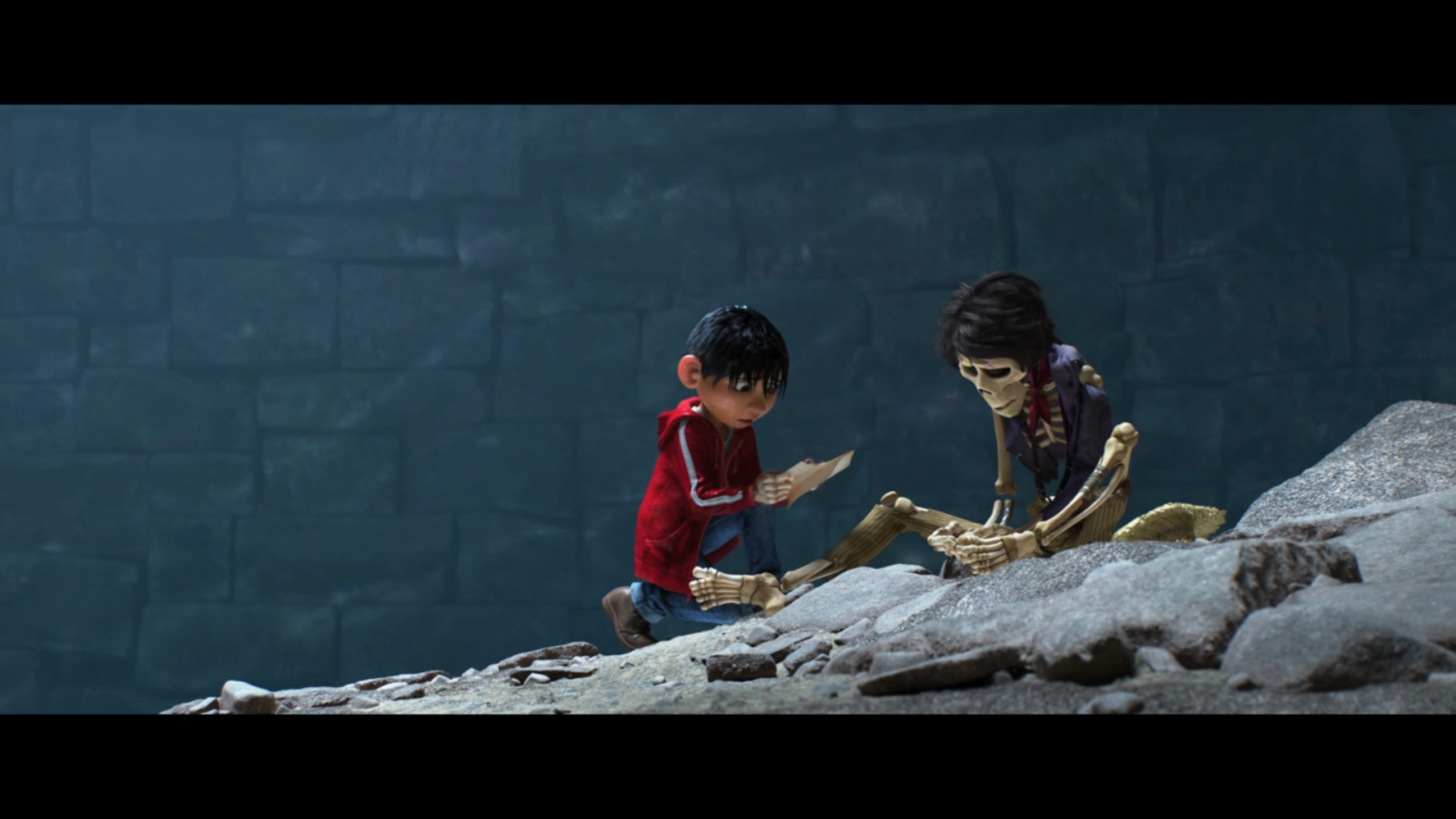 Boy in a red jacket kneels on rocks showing a note to a skeletal figure against a dark stone wall