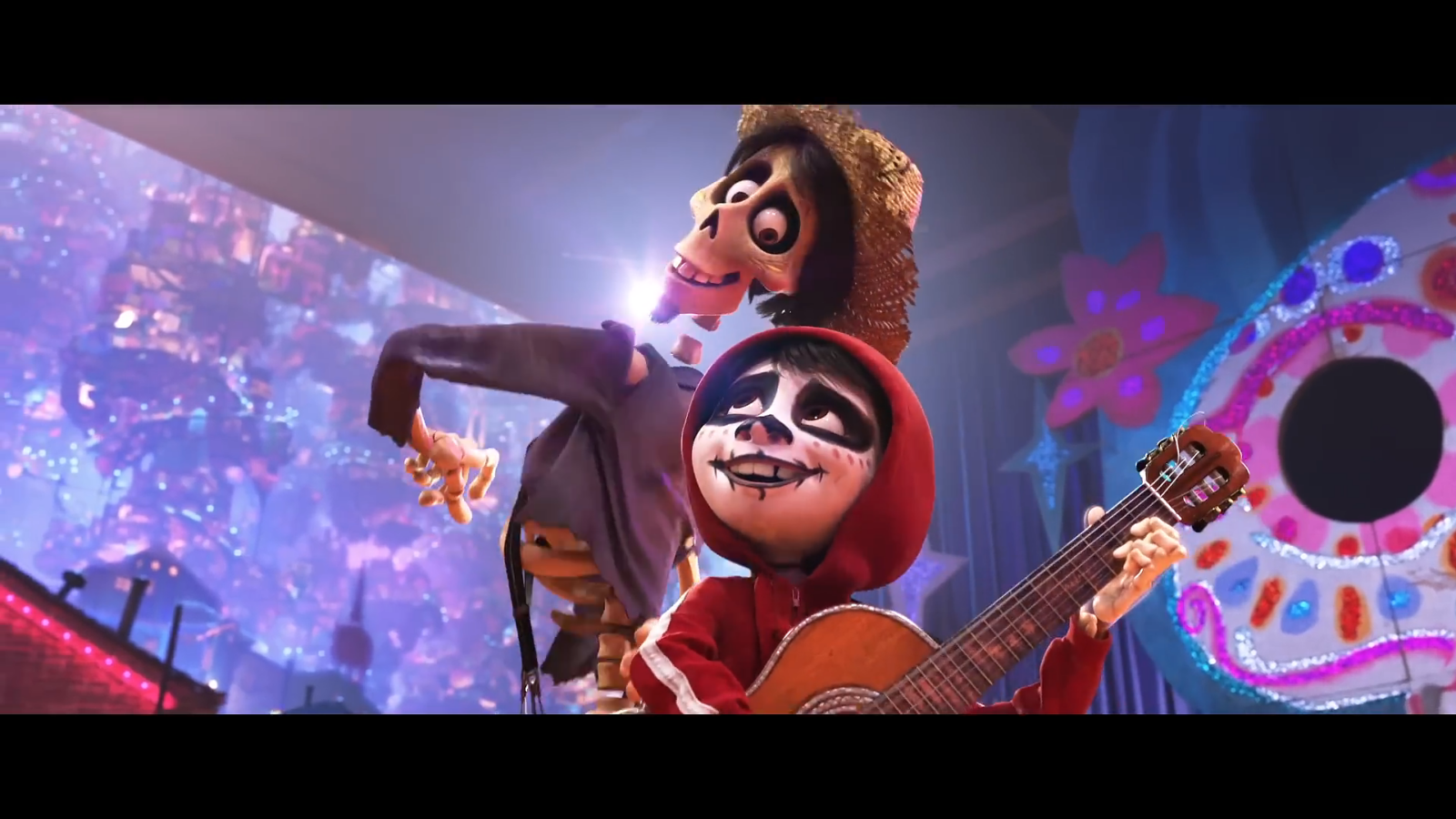 Two performers on a festive stage in Día de los Muertos style masks one in a red hoodie playing a guitar the other a tall skeleton in a straw hat beside him