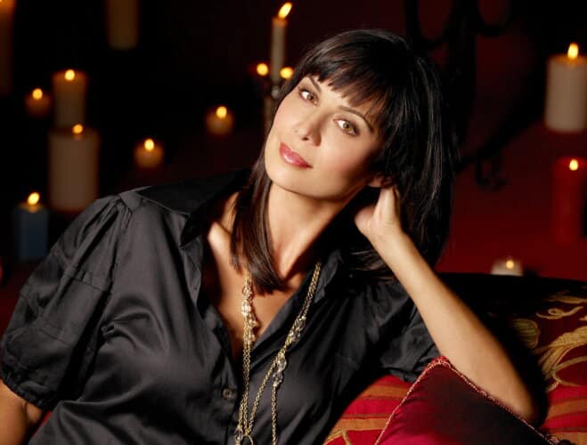 Woman with dark hair in a black satin blouse leaning on her hand on a red couch candlelit background