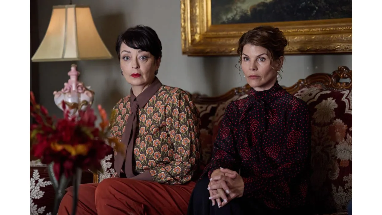 gsm2 Fantasy Land News Two women sit side by side on an ornate patterned sofa wearing vintage blouses and looking solemn in a dimly lit living room