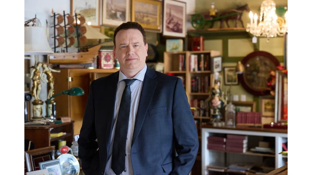gsm5 Fantasy Land News Smiling man in a navy suit and tie stands in an antique shop filled with shelves frames and vintage decor