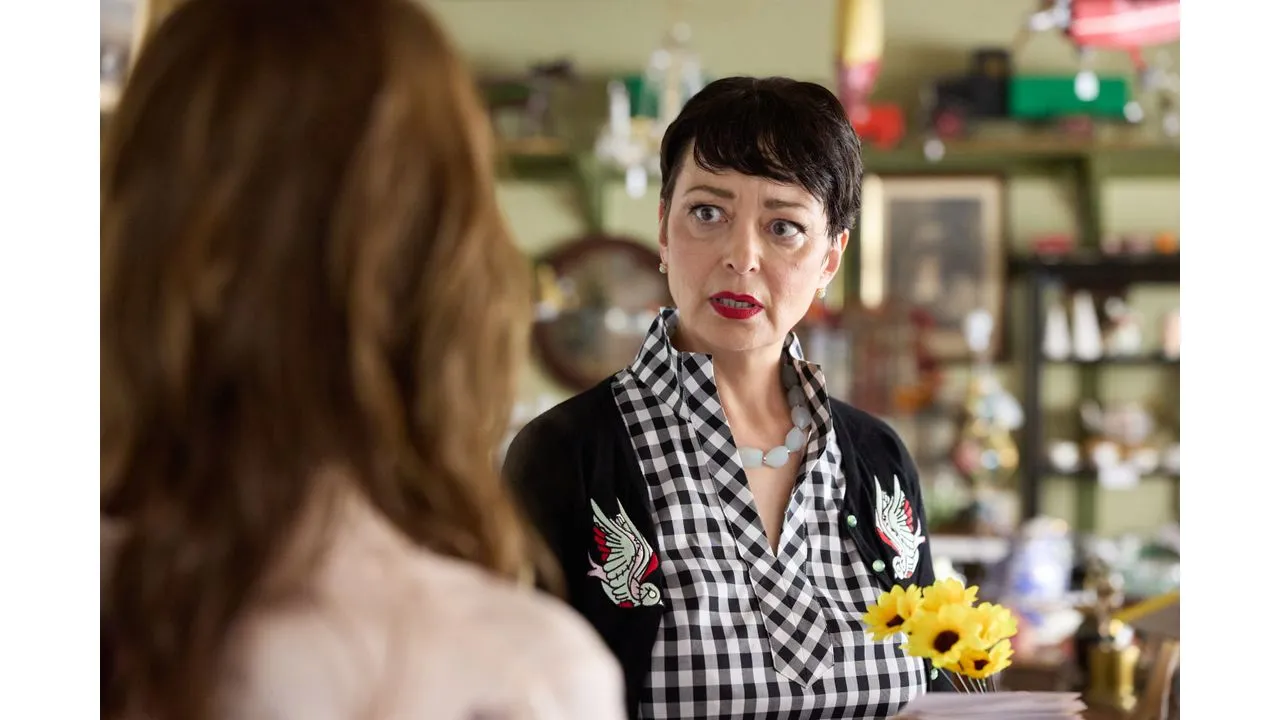 gsmc Fantasy Land News Woman with short black hair and red lipstick speaks to a customer in a shop wearing a black cardigan over a black and white gingham blouse with bird embroidery