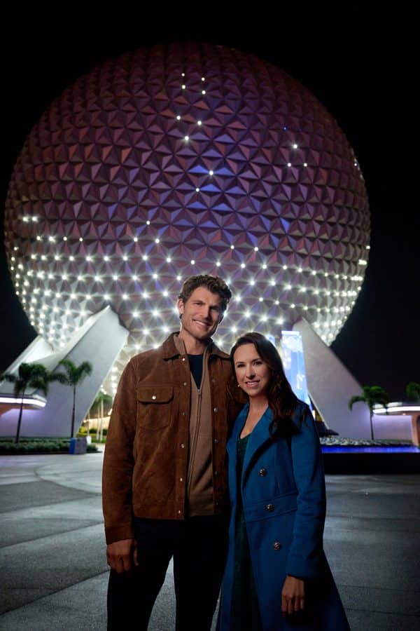Couple posing in front of Epcots Spaceship Earth at night smiling wearing jackets in front of the lit geodesic sphere