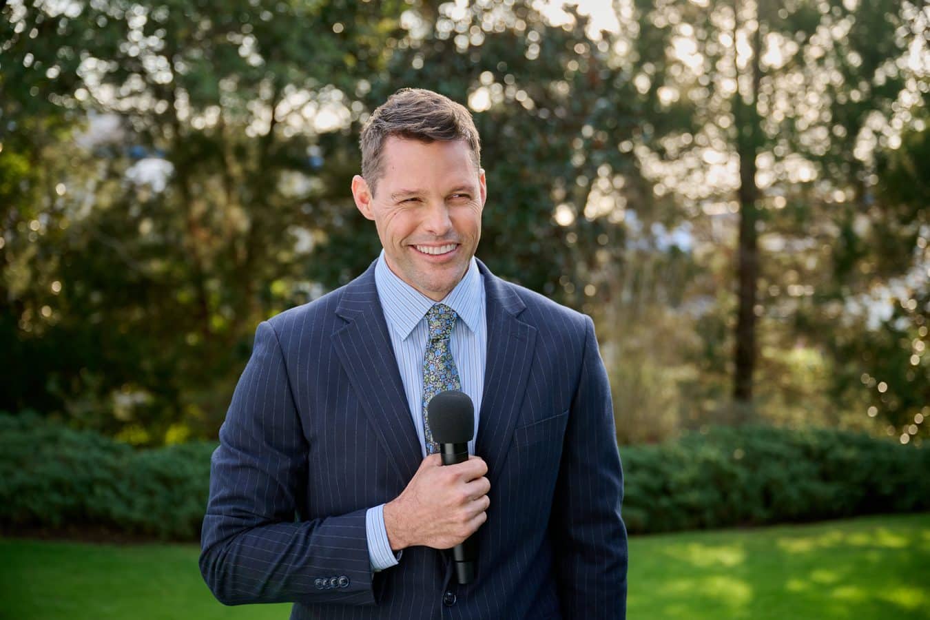 Man in a navy pinstripe suit holding a handheld microphone outdoors smiling at the camera in a park setting
