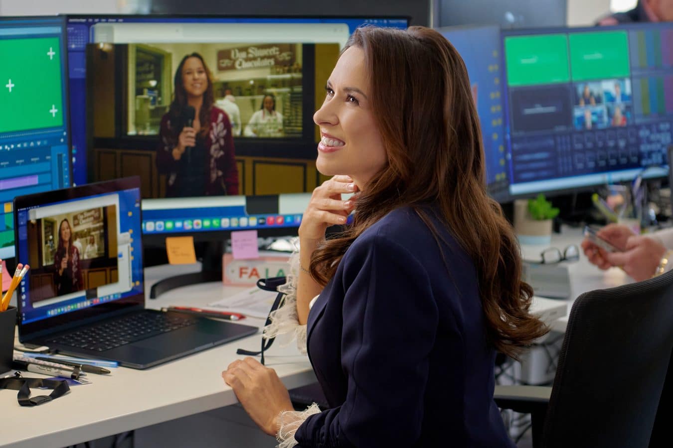 Smiling woman in a navy blazer sits at a desk with several computer monitors in a control room setting