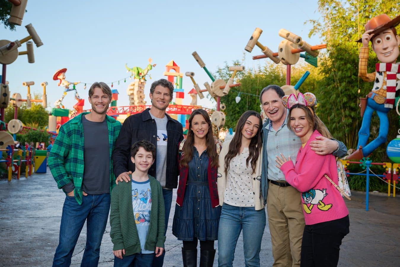 Group of smiling adults and children posing for a photo at a colorful outdoor amusement park with oversized wooden sculptures in the background and bright decorations