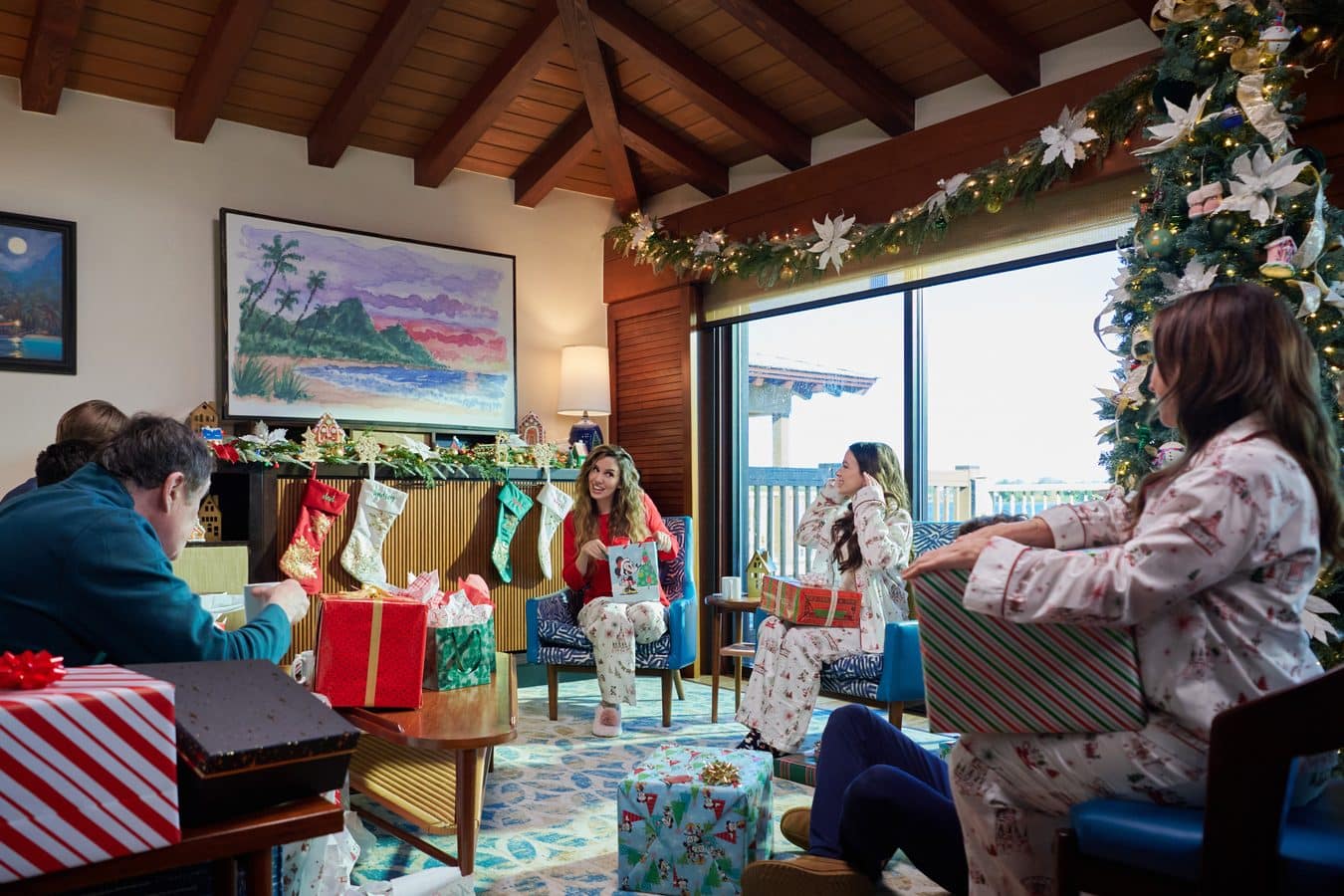 People in festive pajamas sit around a Christmas party room filled with wrapped gifts stockings and a decorated tree