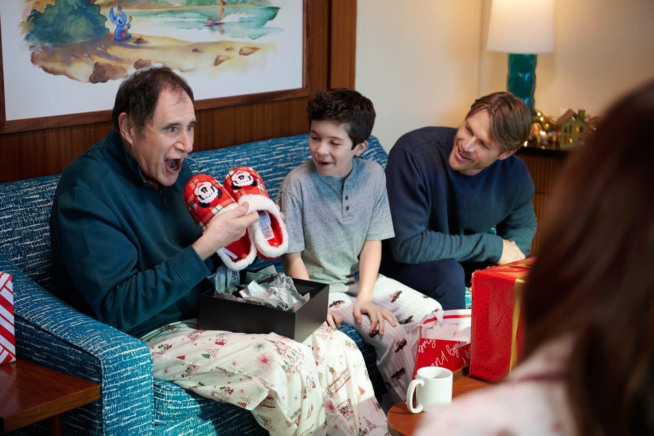 Family members on a blue couch opening Christmas gifts the man on the left holds red plaid Santa slippers while a boy and another man laugh nearby