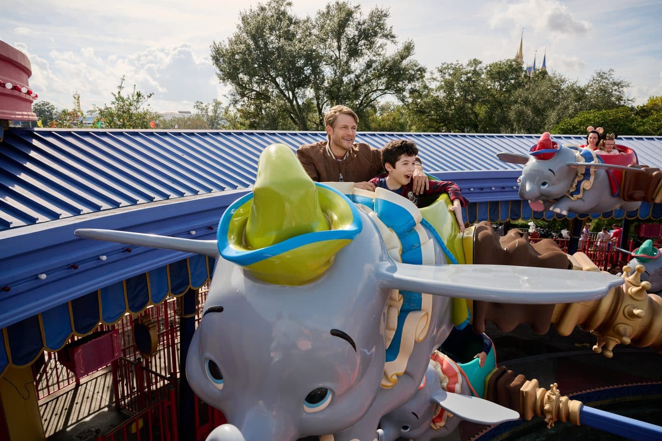 Man and boy ride a colorful Dumbo elephant ride at an amusement park with two girls in Minnie ears in the background