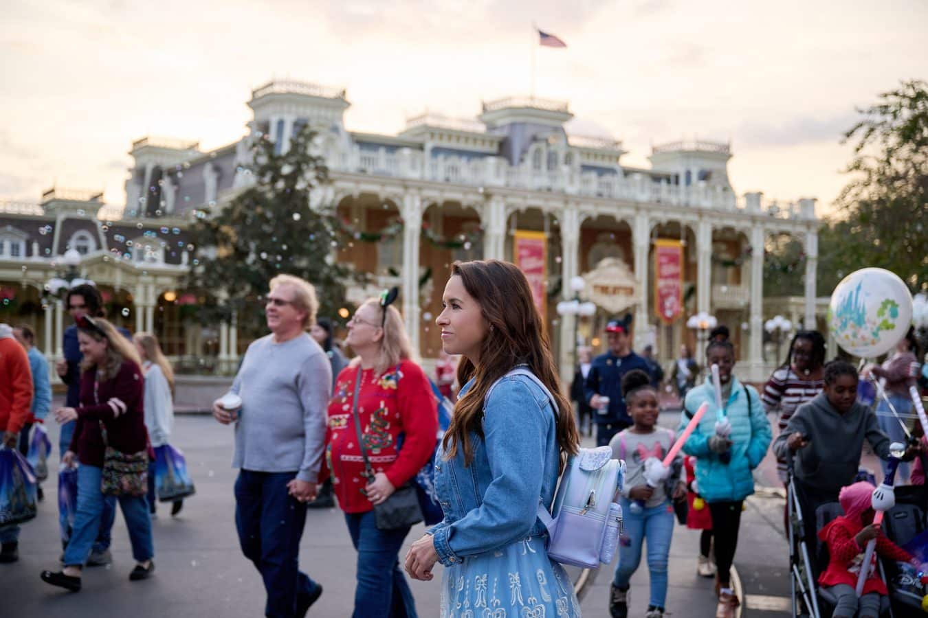 Crowd of visitors in a theme park plaza with a woman in a blue dress in the foreground and an ornate white building in the background