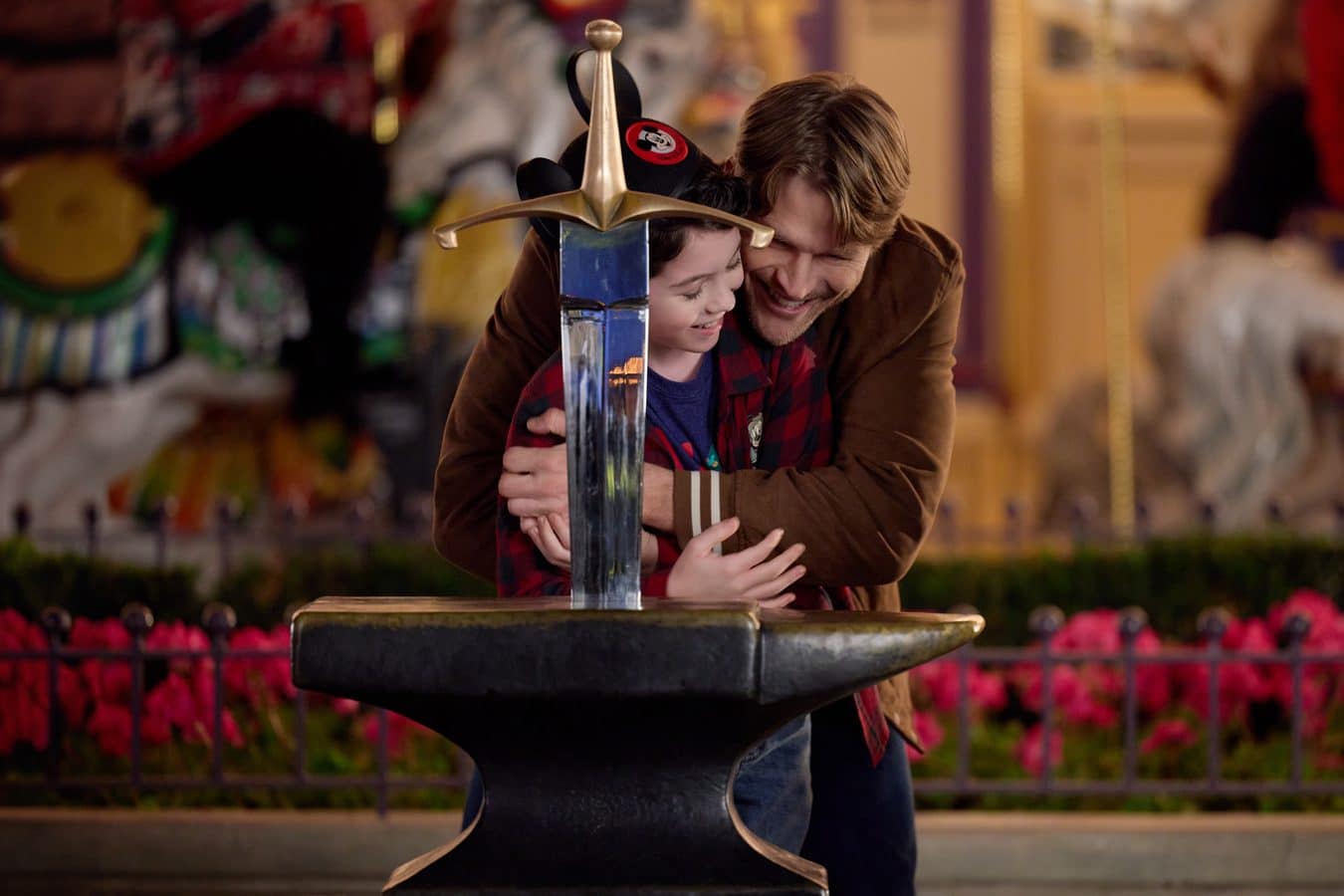 Father and son hug beside a large ceremonial sword embedded in a stone at a colorful outdoor venue