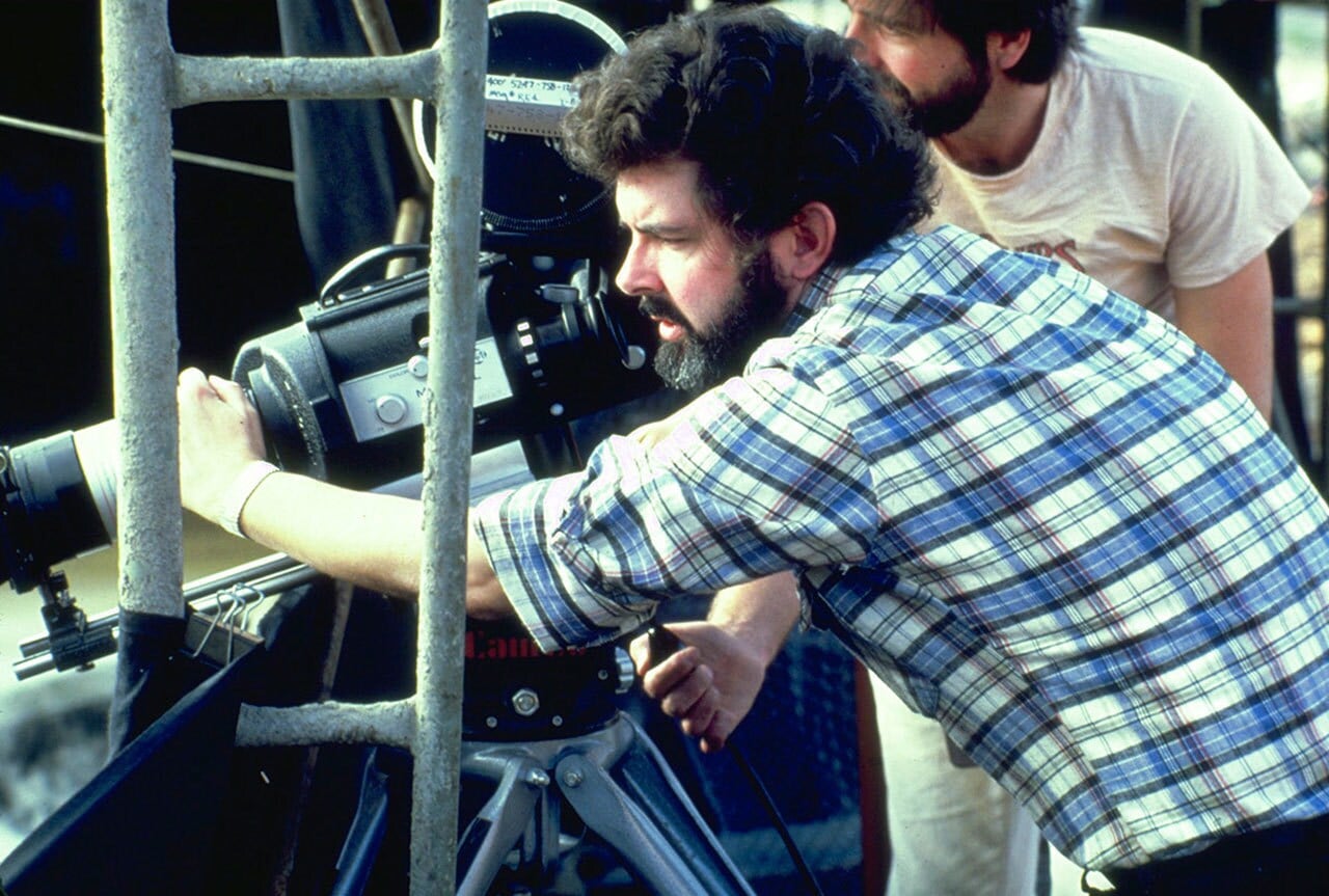 Cinematographer in a plaid shirt adjusts a professional film camera on a tripod behind a metal railing