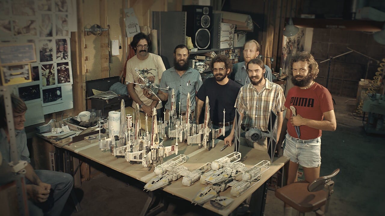 Six men stand around a table filled with model rockets in a workshop setting posing for the photo