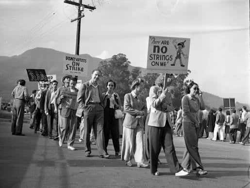 Black and white photo of a labor protest marchers walk along a road holding signs like Disney Artists on Strike and There are no strings on me with hills in the background