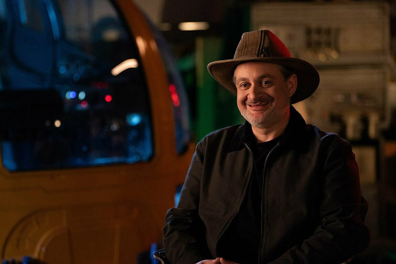 Smiling man wearing a wide brimmed hat and dark jacket sits in a warmly lit room likely a bar or cafe