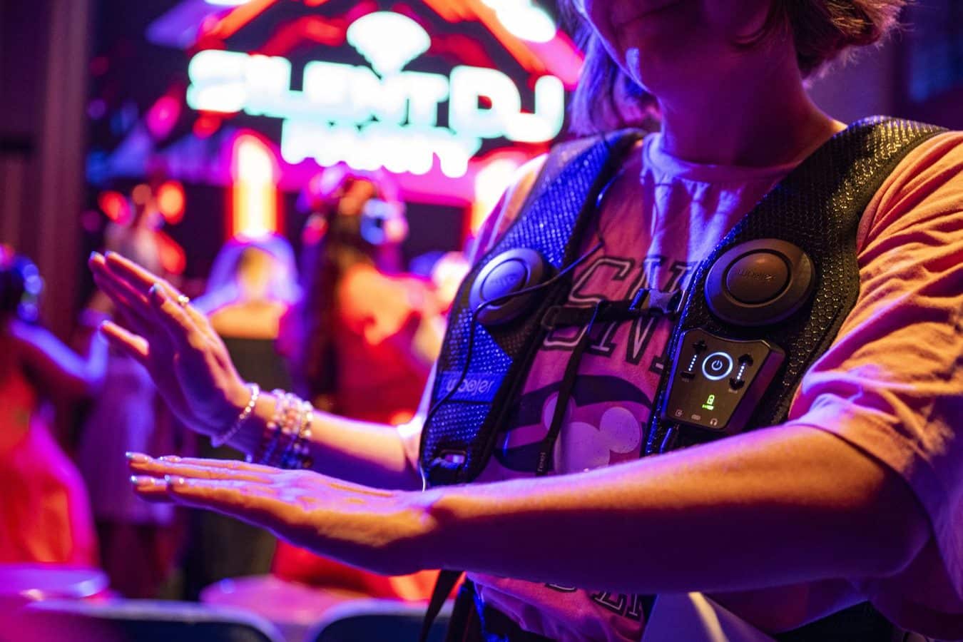 Performer in a sparkly vest claps hands on a neon lit stage with a blurred crowd in the background nearby