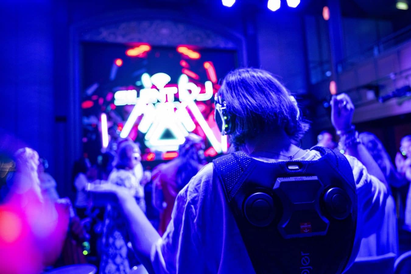 Person seen from behind wearing large headphones on a neon lit club stage with a crowd in the background
