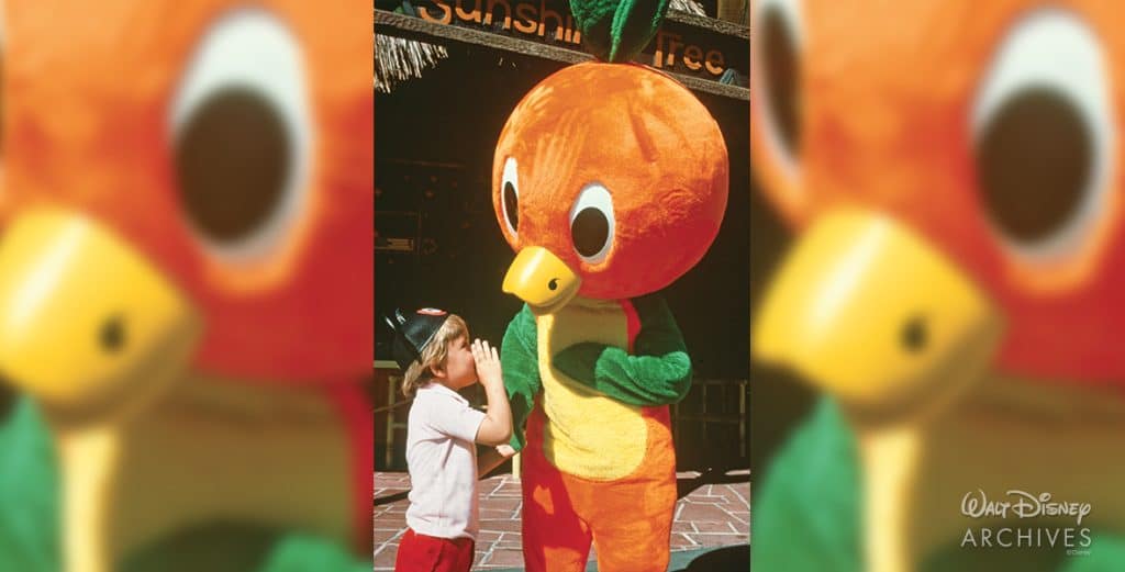 Young girl high fives a large orange parrot mascot outdoors in a Disney park setting