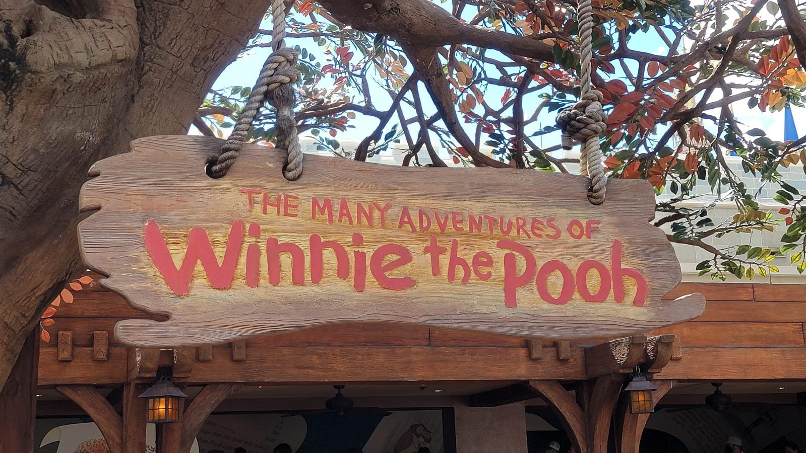 Wooden hanging sign reading The Many Adventures of Winnie the Pooh suspended by thick ropes from a tree with autumn leaves and a wooden storefront in the background to suggest an attraction entrance