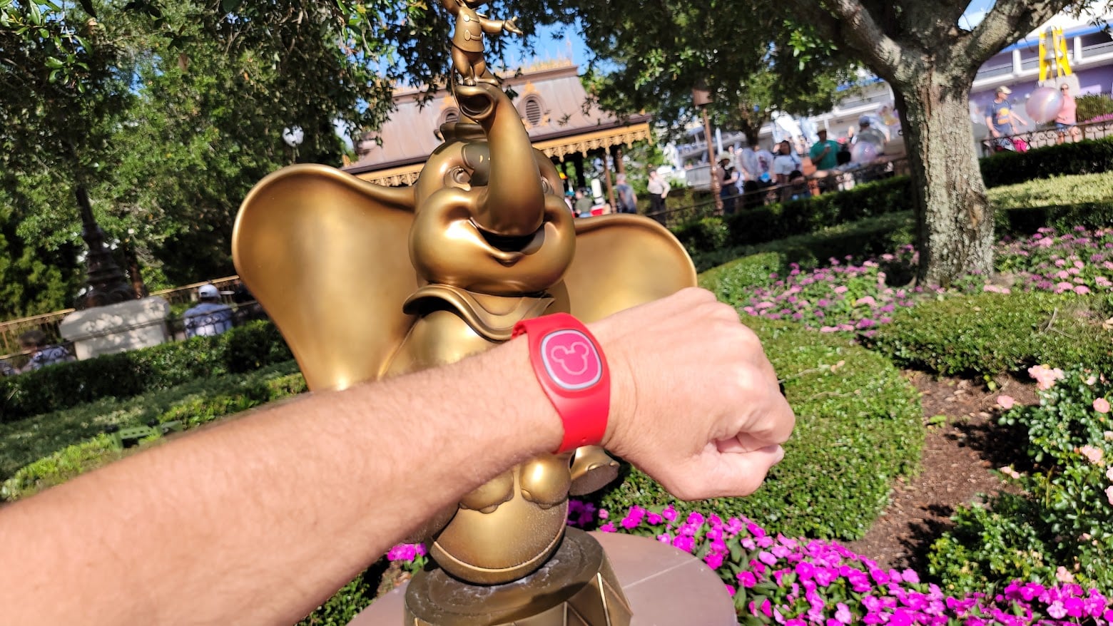Red Disney bracelet on forearm held up beside a shiny gold statue in a Disneyland garden