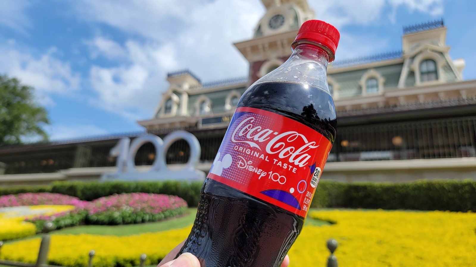 Close up of a Coca Cola bottle held up in front of a Disney park building with colorful flower beds in the foreground and a blue sky behind