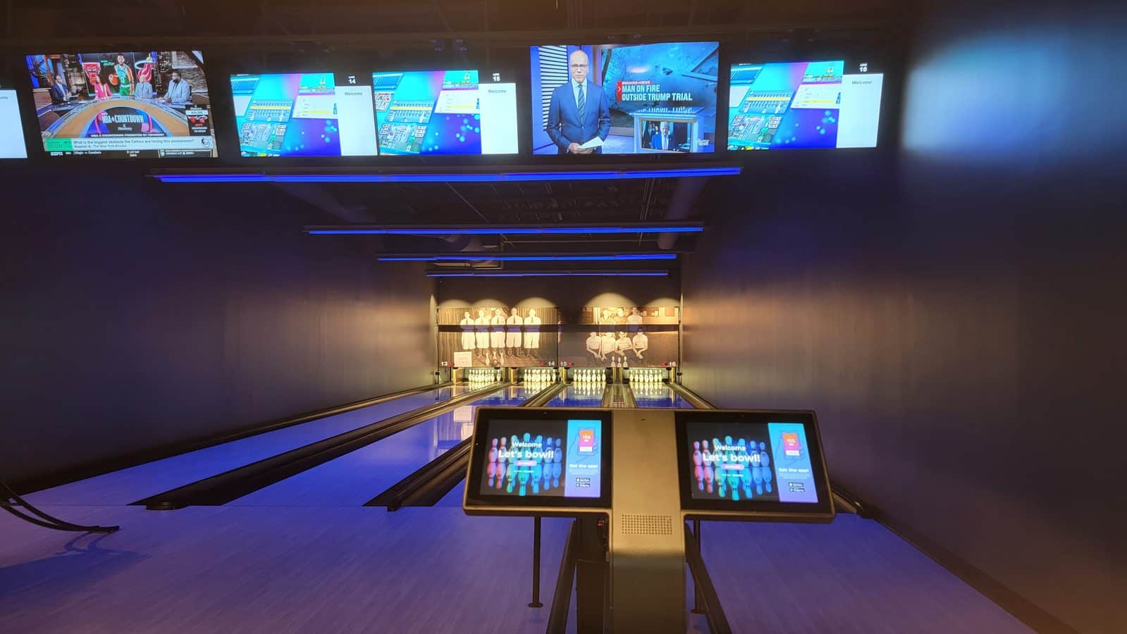 Bowling alley with multiple overhead screens displaying graphics a lane extending toward the pins and a touchscreen console in the foreground