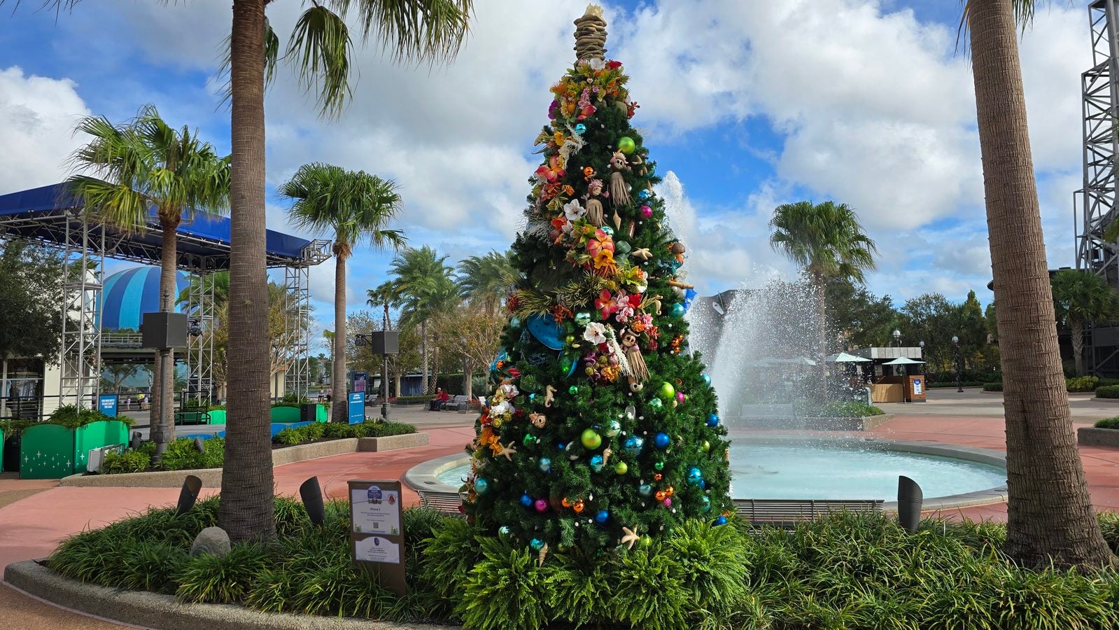 Large Christmas tree decorated with colorful ornaments and flowers next to a fountain in a palm lined plaza