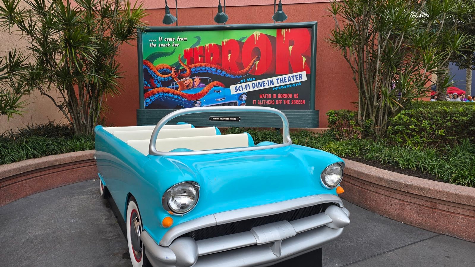 Vintage turquoise car in the foreground with a colorful horror themed sign reading Horror at the Sci Fi Dine In Theater behind it in a theme park setting