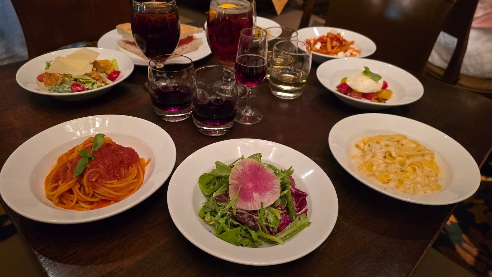 Table set with multiple white bowls of pasta a green salad with radish and creamy pasta plus several glasses of red wine on a dark wooden table